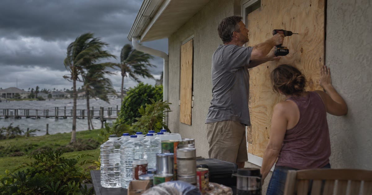 Preparing a home for a hurricane by boarding a window, with stacked water jugs and canned food